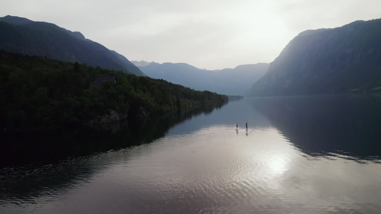 Drone shot over Lake Bohinj in Slovenia as a couple does stand up paddling with mountains in the backdrop as the sun sets