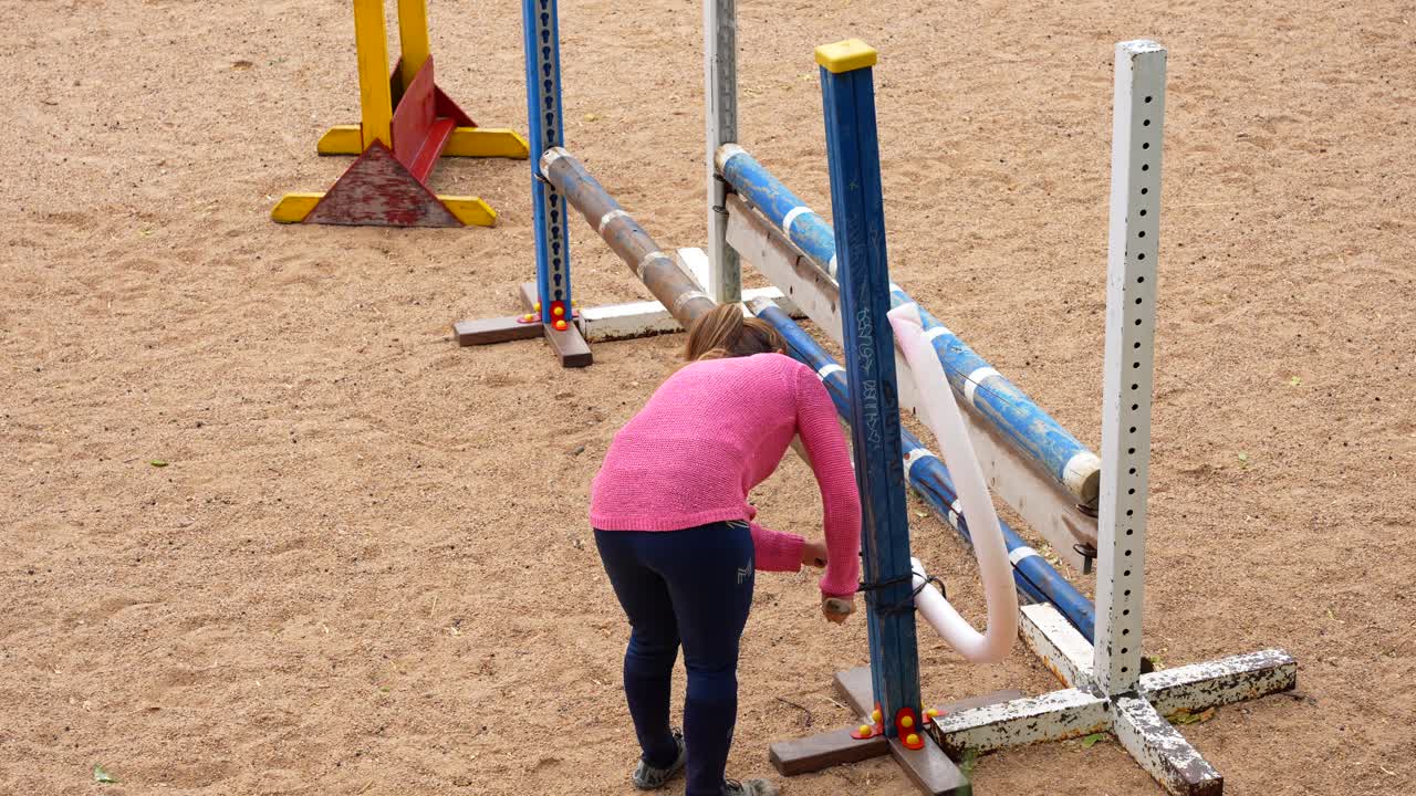 Woman works on a sandy riding track, repairing or adjusting a colorful show jumping hurdle in preparation for an equestrian competition or training session