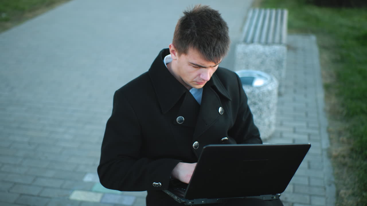 실버 버튼이 있는 검은색 슈트를 입은 데이터 분석가 노트북에서 야외에서 타이핑하고, 배경에는 녹색 잔디, 포장된 산책로, 그리고 벤치 구조가 포함되어 있습니다.