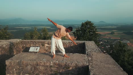 drone push-in shot of indian man stretching and doing yoga pose on top of the hill at sunrise in traditional yogi clothes