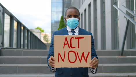 portrait of african american elegant young man in facial mask showing act now" signboard in the street"