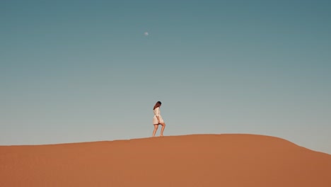 young caucasian woman walking on top of a desert dune during sunset with moon in the background in zagora, morocco