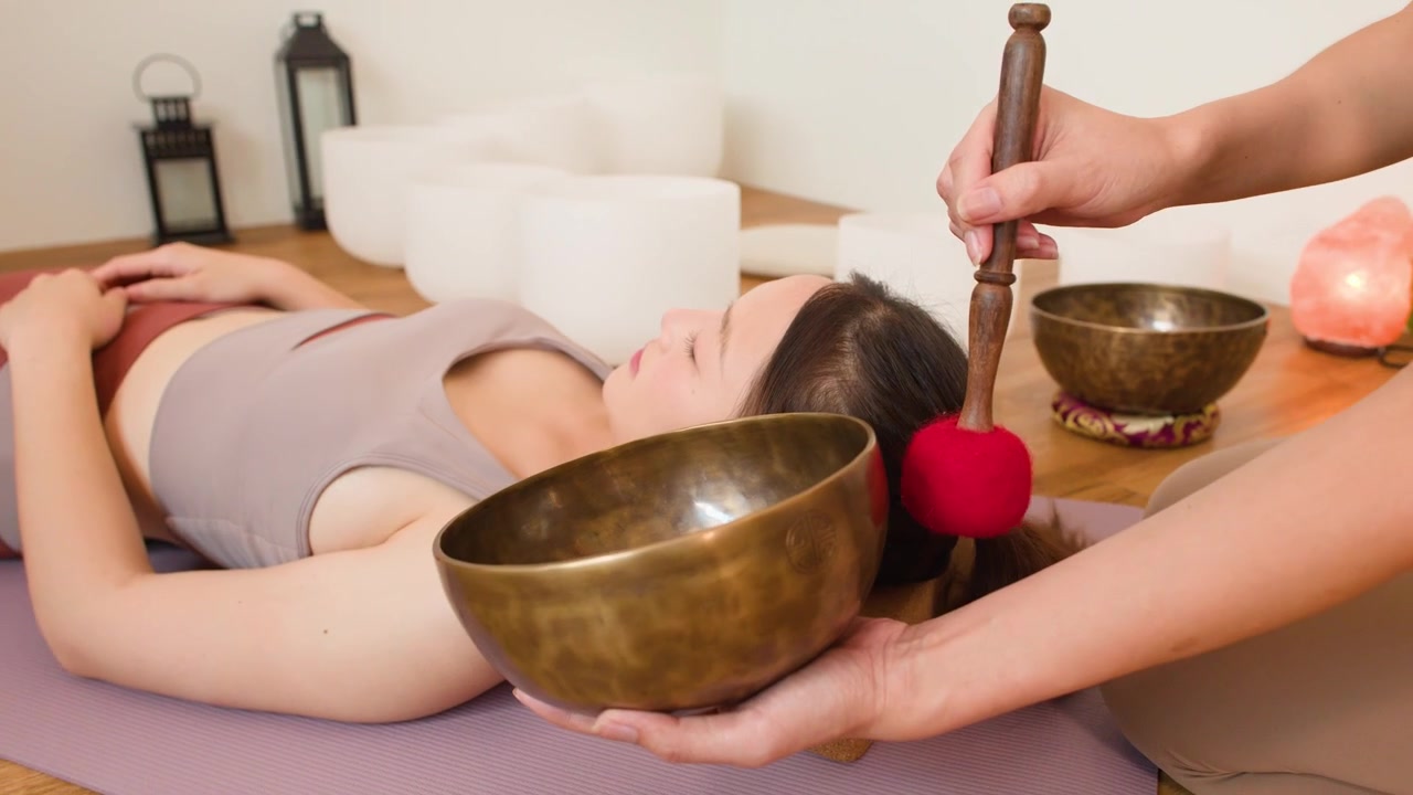 Una donna è sdraiata su un tappetino da yoga mentre un terapista esegue una terapia del suono con ciotole.