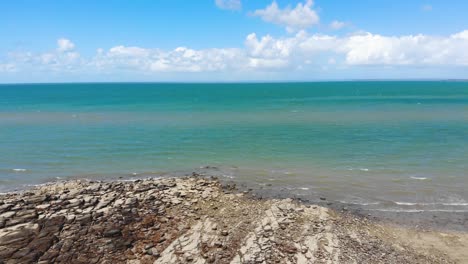 Fly-over-a-beach-made-of-stone,-sand-and-small-rocks-towards-the-gorgeously-tropcial,-artic-blue-and-emerald-green-ocean-sitting-under-a-beautifully-sunny-sky