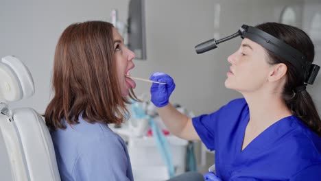 close-up of a confident young brunette girl at an appointment with an ent doctor in a modern clinic. a brunette doctor girl in a blue uniform puts a special stick in the patients mouth to check the throat