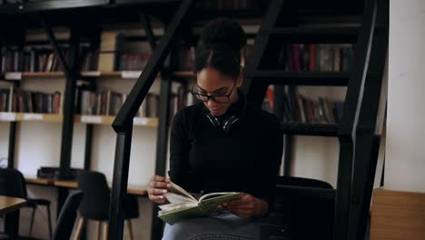 Estudiante-Inteligente-Recopilando-Información-De-Diferentes-Libros,-Chica-Elegante-Con-Suéter-Negro-Y-Auriculares-Alrededor-Del-Cuello-Recopilando-Datos-Para-Los-Libros,-Interior-En-Una-Biblioteca-Moderna