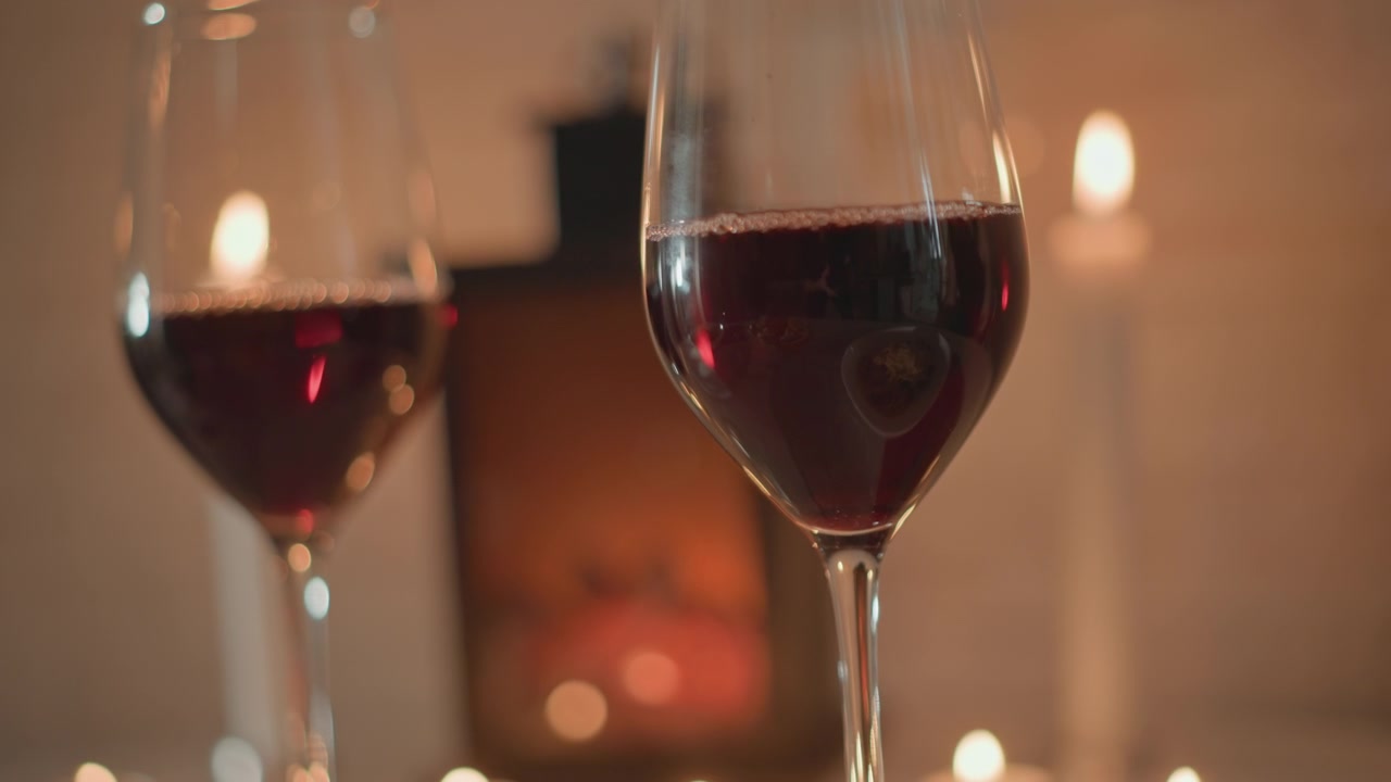 Copo de vinho tinto em primeiro plano mostra líquido suavemente girando e menisco inclinado, enquanto o segundo copo permanece imóvel; a luz quente de velas cria suaves destaques bokeh, superfícies reflexivas e um ambiente de jantar aconchegante.