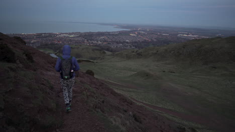 follow tracking shot of girl walking down the arthurs seat mountain on the hiking trail in evening, dusk with city of edinburgh and atlantic ocean in the background