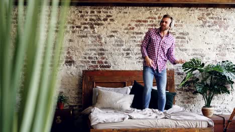Cheerful bearded guy in jeans and shirt is dancing on double bed and singing while listening to music in wireless headphones having fun in bedroom at home.