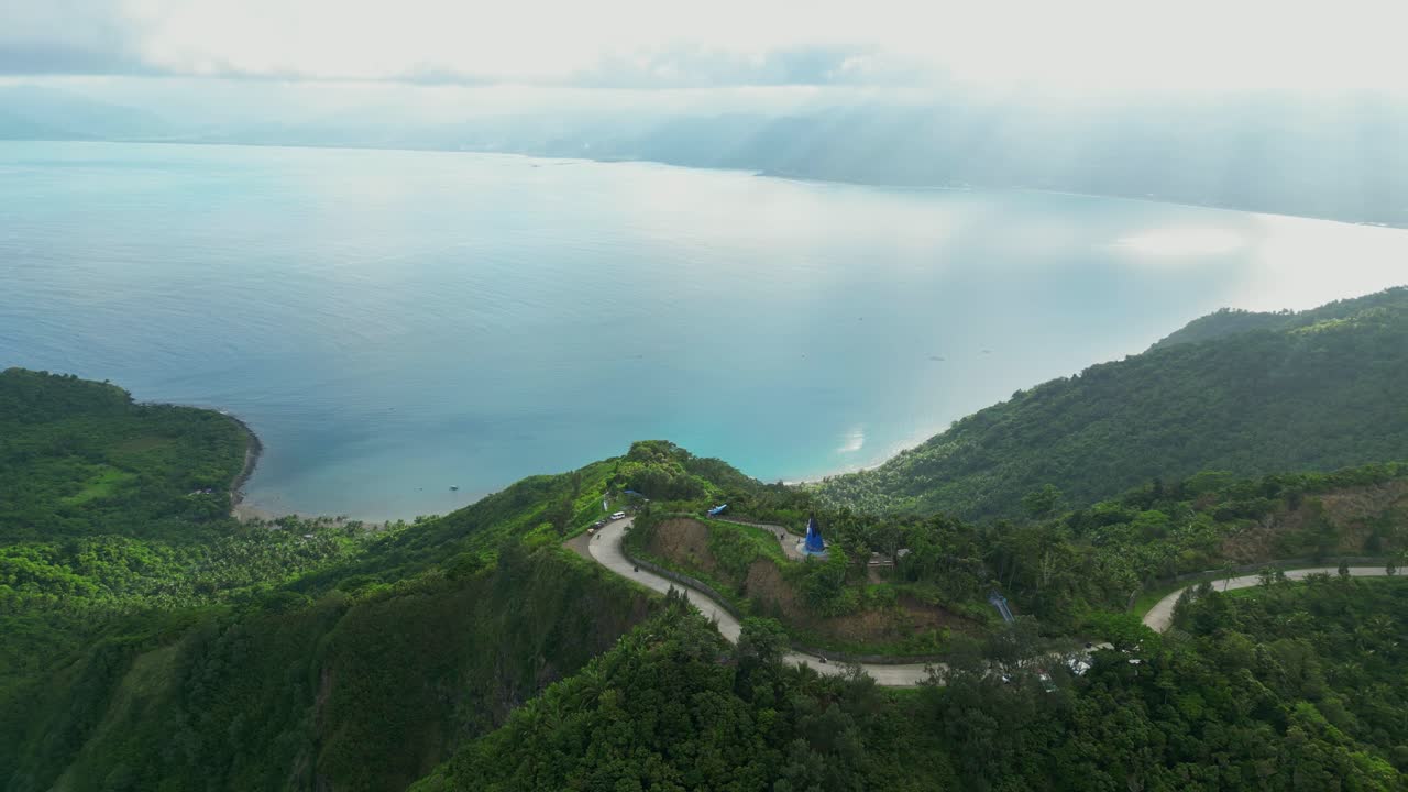 Distant aerial of Dingalan, Aurora, framing La Nuestra Señora dela Paz y Buen Viaje Statue on the hilltop as golden sun rays break through clouds and illuminate the sea below