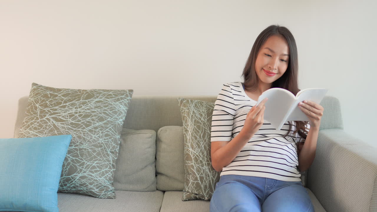 젊은 성인 아시아 여성의 우아한 평상복은 화려한 베개와 함께 소파에 앉아 빈 흰색 저널 책을 읽고 현대적인 실내 홈 호텔 환경과 복사 공간을 갖추고 있습니다.