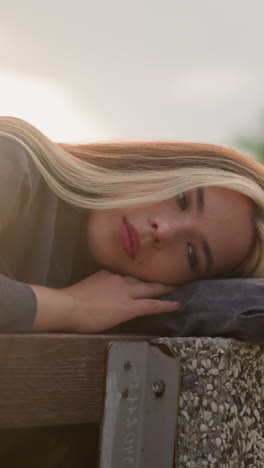 mujer sentada en el borde de un banco descansando la cabeza sobre el brazo en un parque al aire libre sereno con la luz del sol suave reflejándose a su alrededor, el fondo incluye árboles y un edificio