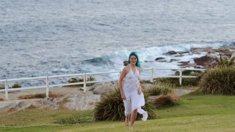 Runaway-bride-runs-home,-barefoot-blue-hair-white-dress-runs-up-grass-bank-from-ocean,-SLOMO