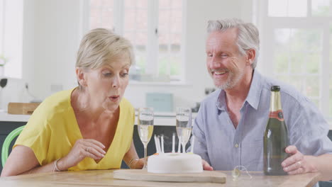 Senior-Woman-Blowing-Out-Candles-On-Cake-Celebrating-Birthday-With-Glass-Of-Champagne-With-Husband