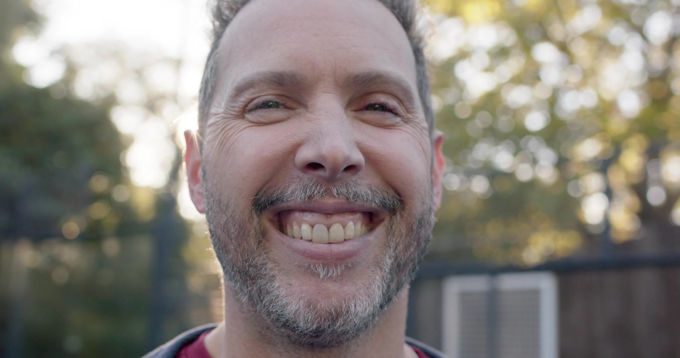 Middle-aged man in maroon tee smiling in backyard by wood fence, lens flare causing smile widening