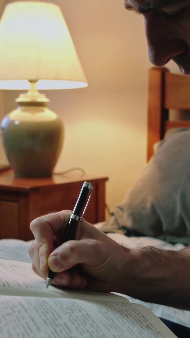 Video verticale: Uomo appoggiato con camicia grigia che porta una penna stilografica a un libro aperto sul letto, prendendo appunti vicino a una lampada.