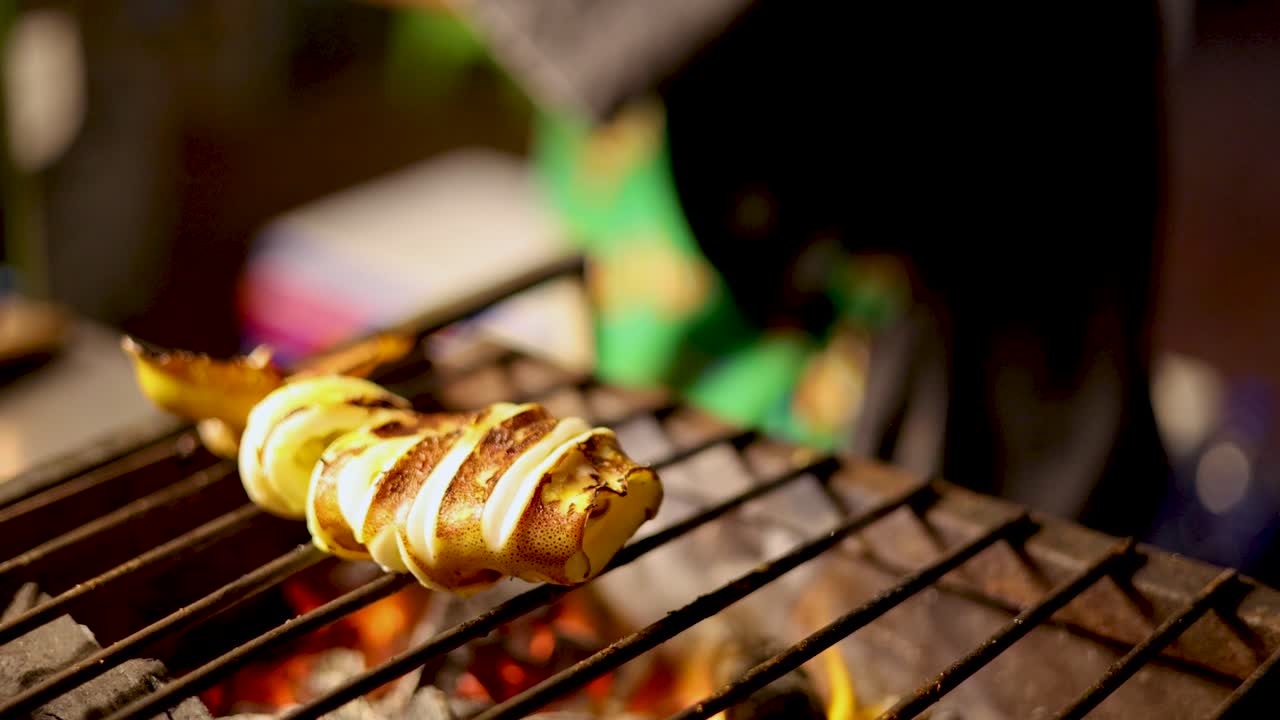 Vendor grills whole squid on skewers over charcoal, vibrant street market, warm night lighting