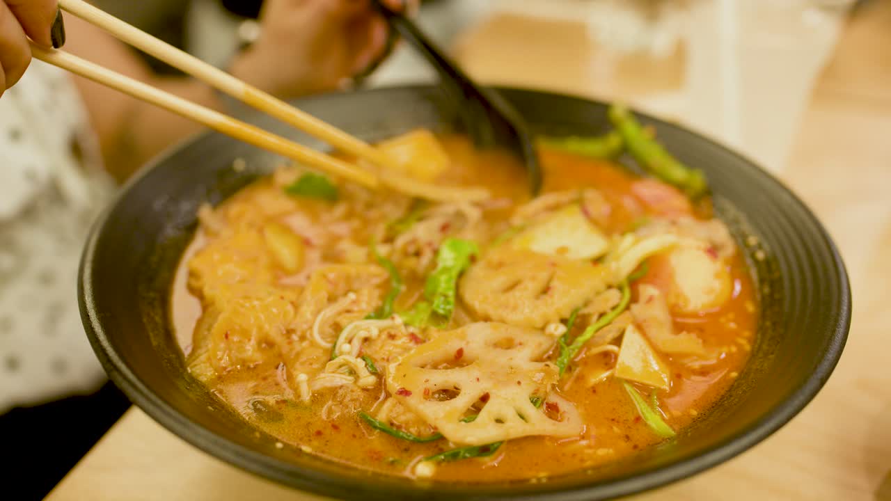 Hand uses chopsticks to pick up lotus root from hot, vibrant Asian noodle soup bowl