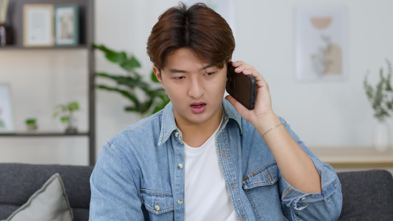 Một chàng trai trẻ người châu Á ngồi trên диван, trò chuyện điện thoại thông minh với vẻ mặt bực bội