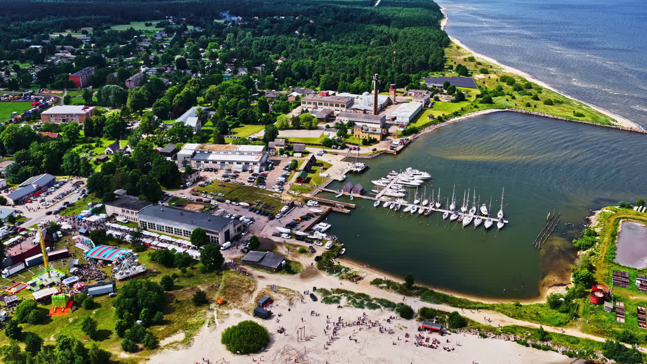 A wide aerial view captures the summer Zvejnieksvētki (Fisherman's Festival) in the coastal town of Engure, Latvia, showing the harbor full of sailboats and a bustling fair