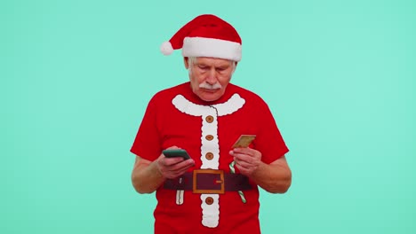 Älterer-Weihnachtsgroßvater-Mit-Kreditkarte-Und-Smartphone,-Der-Geld-Beim-Online-Einkauf-überweist