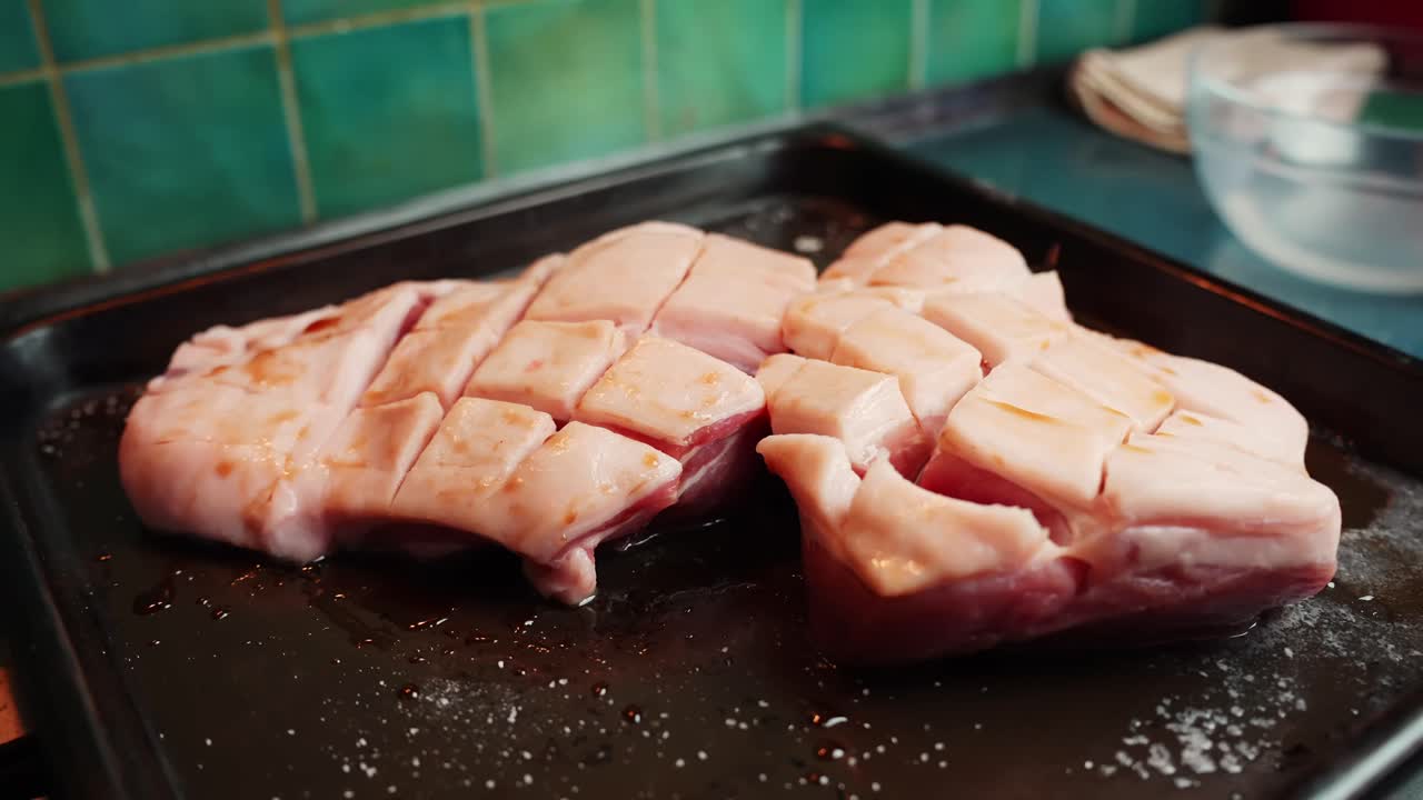 Plano macro de uma mão espalhando temperos secos sobre barriga de porco marcada numa assadeira, preparando carne crua para assar numa cozinha doméstica