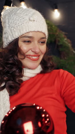 beautiful lady with large red christmas balls. happy young woman in white knitted scarf and hat holds christmas baubles in nicely decorated room in new year. vertical video