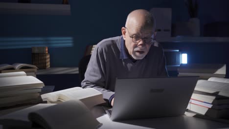elderly researcher or historian man reading a book, using laptop alone in dark study room.