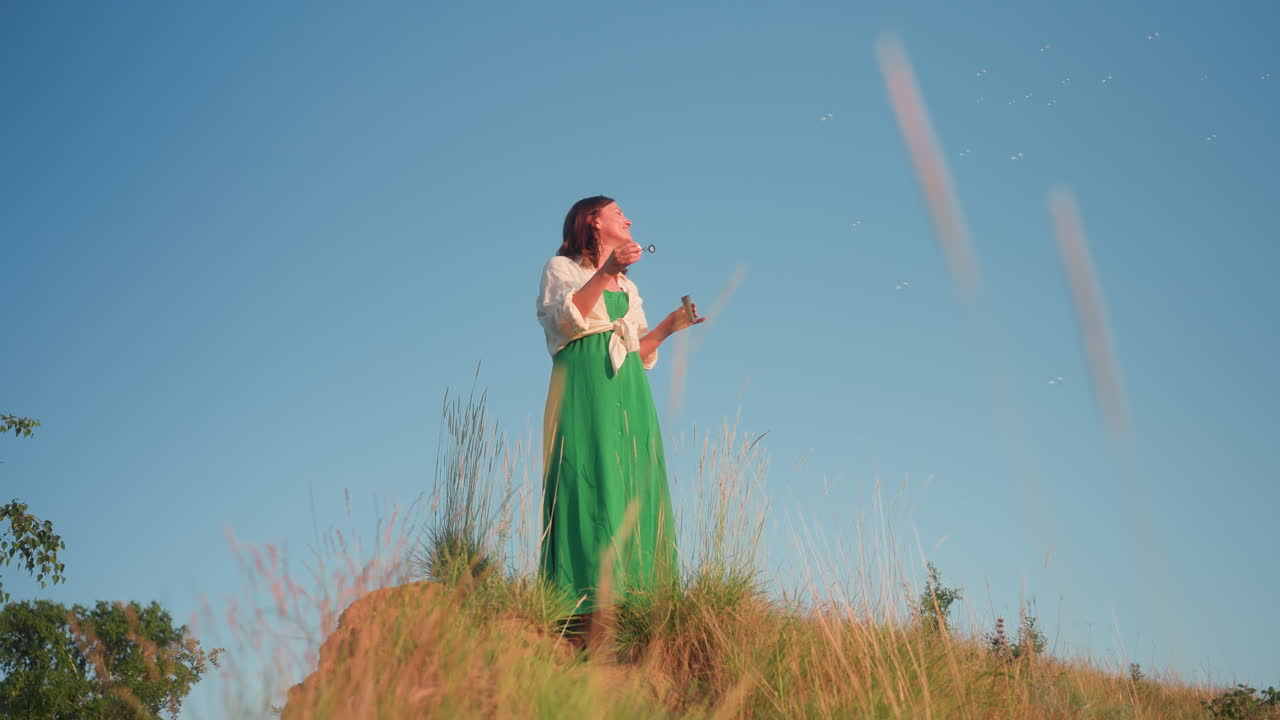 Woman standing near cliff edge blowing soapy bubbles under clear blue sky on grassy hilltop, green dress flowing, gentle breeze stirring grasses as she enjoys serene