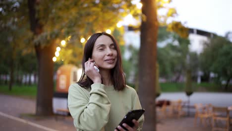 una chica morena en un suéter verde escucha música en los auriculares contra el telón de fondo de un parque nocturno. luces brillantes cerca