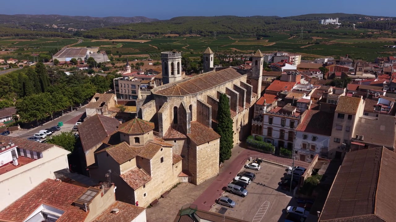 Centro città di Sant Julia de l'Arboc in Catalogna, Spagna, con una chiesa medievale in pietra circondata da edifici con tetti in terracotta, vigneti e verdi colline sullo sfondo.