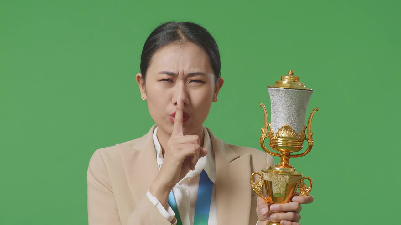 금메달과 트로피를 입은 슈트를 입은 아시아 사업가 여성의 클로즈업은 스튜디오의 녹색 스크린 배경에서 첫 번째 우승자로서 승리하는 비밀이라고 말하는 <shh> 제스처를 만니다.