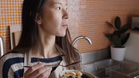 young asian woman eating breakfast in kitchen enjoying healthy oatmeal cereal mixed with fruit relaxing at home on weekend