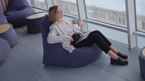 Happy relaxed woman sits in bag armchair with computer on her laps. Dreamy lady stretches hands and puts them behind her head. View from above.
