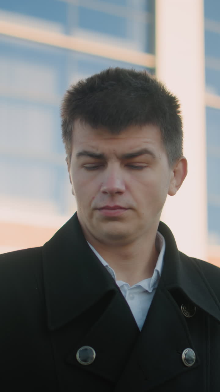 검은색 슈트를 입은 러시아 사업 거장, 동의 표시로 고개를 끄덕이는 동안 전화를 들고, 배경에는 현대적인 유리 건물과 도시 구조물이 있습니다.