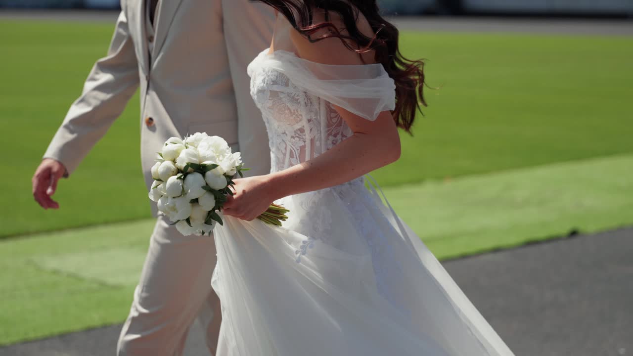 Bride and groom walking together on a sunny day, with the bride holding a bouquet of white roses in her elegant off-shoulder wedding dress