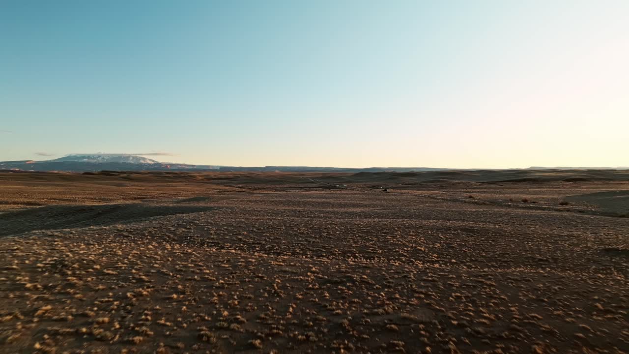 High aerial panoramic establishing approach of an SUV driving across the Moab desert at sunset, glowing golden terrain