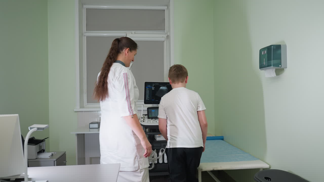 Lab scientist greets young boy with warm smile inside clinic, gestures toward patient bed while engaging in friendly interaction, standing near ultrasound machine during medical consultation setup