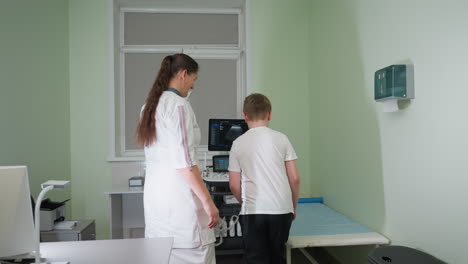 lab scientist greets young boy with warm smile inside clinic, gestures toward patient bed while engaging in friendly interaction, standing near ultrasound machine during medical consultation setup