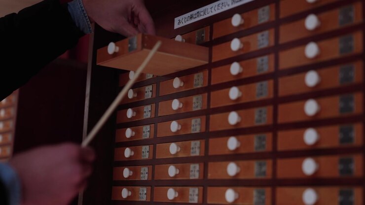 Mani che estraggono un bastoncino della fortuna da un mobiletto in legno con cassetti numerati per omikuji in un santuario giapponese.