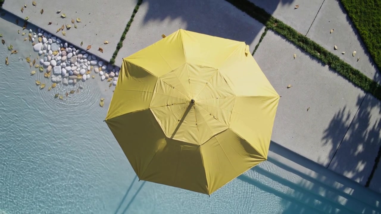La telecamera si allontana rivelando un ombrellone giallo aperto centrato sopra una piscina, mostrando ciottoli e lastre di pavimentazione.
