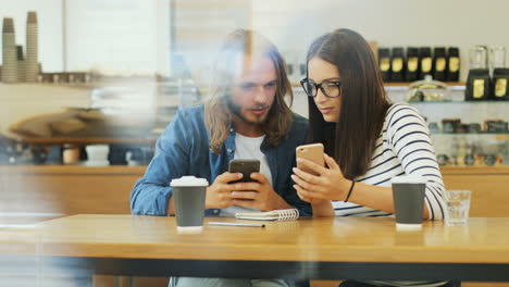 caucasian man and woman friends using smartphone and talking while they are sitting at a table in a cafe