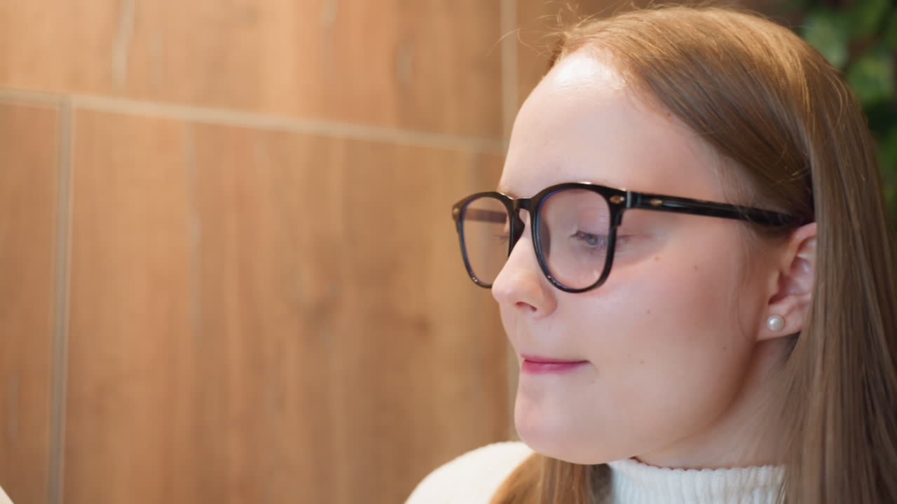 close up of young woman in glasses and white turtleneck sweater applying lipstick while looking slightly upward in warmly lit indoor setting with wooden wall and green plants in soft background