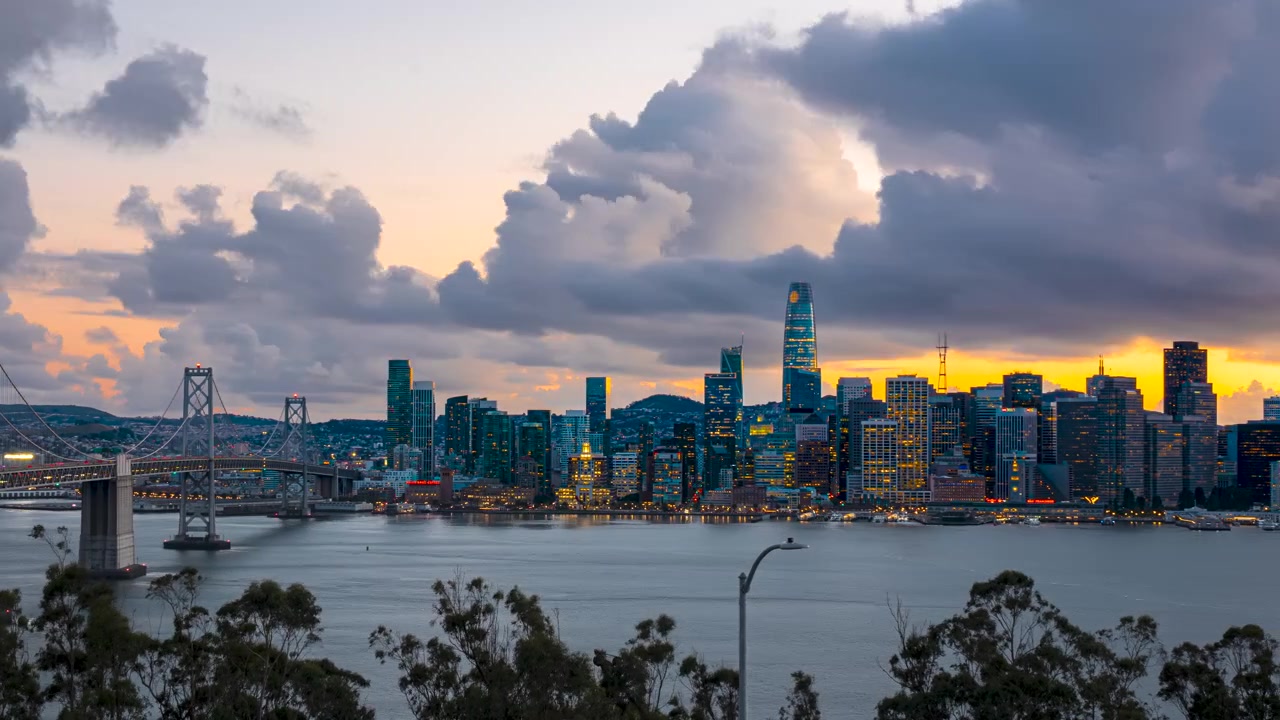 Timelapse estático panorâmico do horizonte de São Francisco e da Ponte da Baía enquanto o céu fica laranja ao pôr do sol.