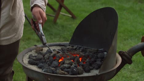 a revolutionary war reinacter blacksmith puts a piece of metal in hot coals to heat up while he pumps the bellows