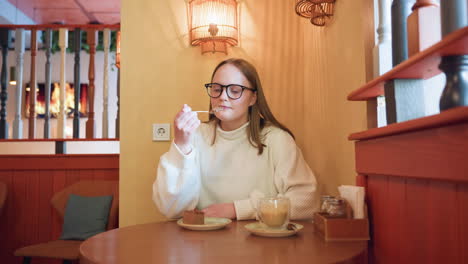 woman seated at round table in cafe, gently savoring chocolate cake , glass of latte beside her, background features soft lighting and television displaying animated fireplace flame
