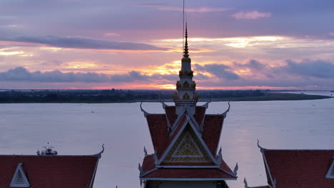 a traditional khmer temple roof and spire silhouette against a vivid river sunset, with boats on the water and the distant horizon glowing in pinks and oranges