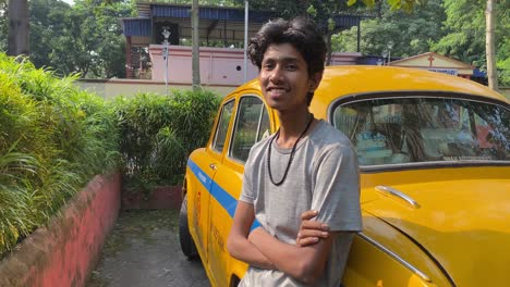 an young indian taxi driver in grey t-shirt standing near his taxi and looking at the camera