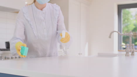 woman in face mask at home in kitchen wearing rubber gloves cleaning down work surface using cleaning spray during health pandemic  - shot in slow motion