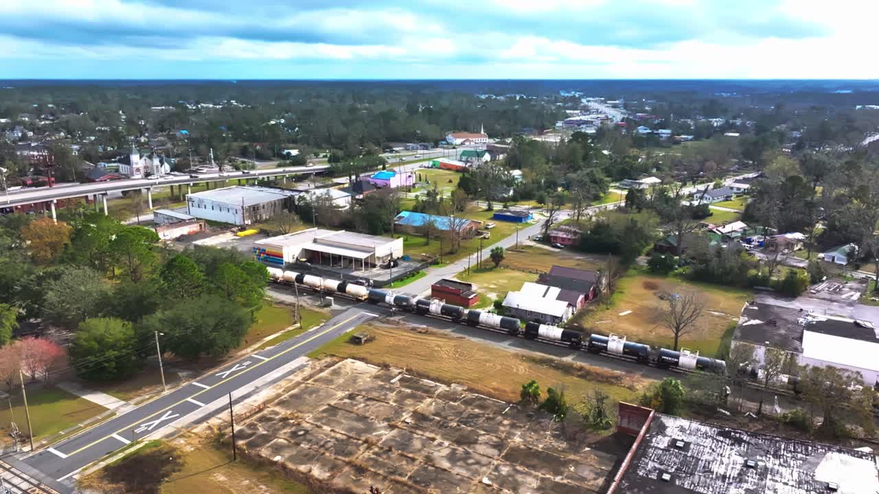 A Freight Train Moves Through a Small Town, Passing by Houses, Commercial Buildings, and a Highway Overpass in the Background - Static Shot